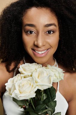Göz kamaştırıcı Afro-Amerikan bir kadın, bej arka planda elinde bir buket beyaz gül tutarken doğal makyajıyla sıcak bir şekilde gülümsüyor..