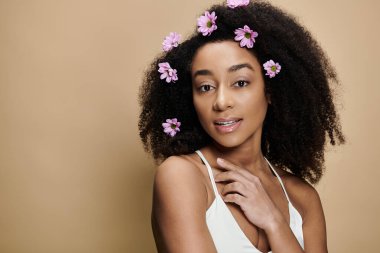 Güzel bir Afro-Amerikalı kadın doğal makyajıyla ve saçlarında çiçeklerle bej bir arka plana hafifçe gülümsüyor..