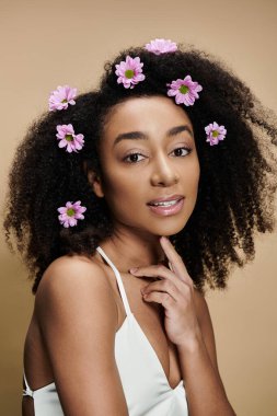 Güzel bir Afro-Amerikalı kadın doğal makyajıyla ve saçlarında çiçeklerle bej bir arka plana hafifçe gülümsüyor..