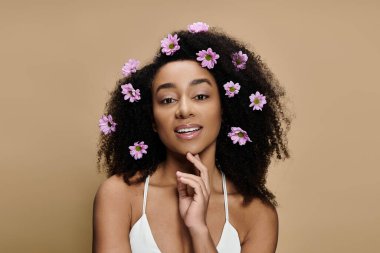 Güzel bir Afro-Amerikalı kadın, doğal makyajı ve saçlarında çiçeklerle bej bir arka plana karşı poz veriyor..