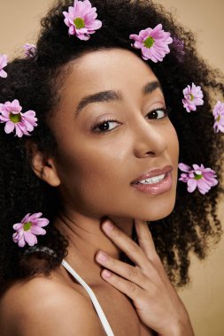 Güzel bir Afro-Amerikalı kadın doğal makyajıyla ve saçlarında çiçeklerle bej bir arka plana hafifçe gülümsüyor..