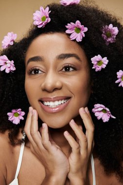 Doğal makyajı ve pembe çiçekleri olan güzel bir Afro-Amerikalı kadın bej bir arka fona gülümsüyor..