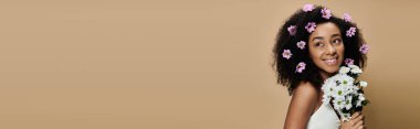 Saçında çiçekler olan Afro-Amerikalı bir kadın bej arka planda parlak bir şekilde gülümsüyor..