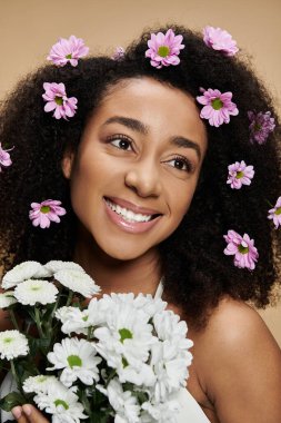 Saçları doğal makyajlı ve çiçekli güzel bir Afro-Amerikalı kadın bej bir arka plana sıcak bir şekilde gülümsüyor..