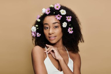 Saçları doğal makyajlı ve çiçekli güzel bir Afro-Amerikalı kadın bej arka planda kameraya hafifçe gülümsüyor..