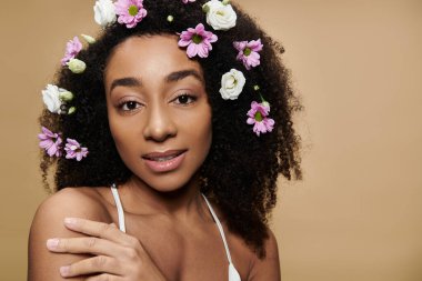 Doğal makyajlı ve saçında çiçekler olan güzel bir Afro-Amerikalı kadın bej arka planda poz veriyor..