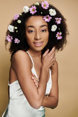 Doğal makyajlı ve saçında çiçekler olan güzel bir Afro-Amerikalı kadın bej arka planda poz veriyor..