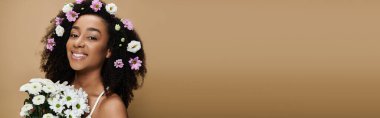 Güzel bir Afro-Amerikalı kadın, doğal makyajı ve saçlarında çiçeklerle bej bir arka plana gülümsüyor..