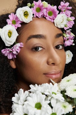 Doğal makyajlı ve saçında çiçekler olan güzel bir Afro-Amerikalı kadın bej arka planda poz veriyor..