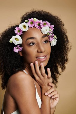 Doğal makyajlı ve saçında çiçekler olan güzel bir Afro-Amerikalı kadın bej arka planda poz veriyor..