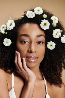 Doğal makyajlı ve saçında çiçekler olan güzel bir Afro-Amerikalı kadın bej arka planda poz veriyor..