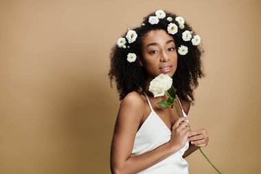 Doğal makyajlı ve saçında çiçekler olan güzel bir Afro-Amerikalı kadın bej arka planda poz veriyor..