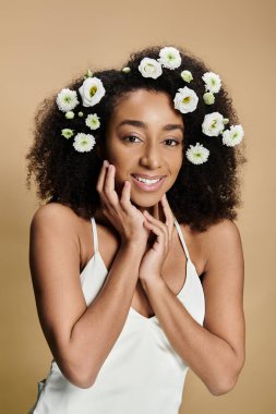 Doğal makyajlı ve saçlarında çiçekler olan güzel bir Afro-Amerikalı kadın bej bir arka plana sıcak bir şekilde gülümsüyor..