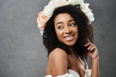 Beyaz bir gelinlik giymiş güzel bir Afro-Amerikalı gelin ve bir çiçek tacı gri bir arka planın önünde dururken gülümsüyor..