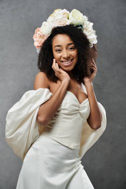 A beautiful African American bride in a white wedding dress with flowers in her hair smiles radiantly against a grey backdrop. clipart