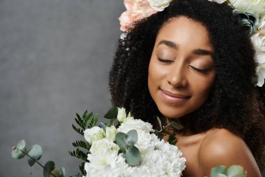 Güzel bir Afro-Amerikan gelini saçlarında çiçeklerle hafifçe gülümser, buketinin kokusundan zevk alırken gözlerini kapatır..