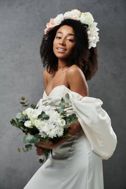 Saçında çiçek tacıyla beyaz gelinlik içinde güzel bir Afrikalı Amerikalı gelin gri bir fonda duruyor..