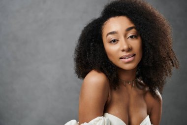 Beyaz gelinlikli güzel bir Afro-Amerikan gelin gri arka planda poz verirken kameraya gülümsüyor..