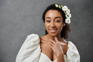 Beyaz bir gelinlik içinde güzel bir Afro-Amerikalı gelin saçlarında çiçeklerle gri bir arka plana gülümsüyor..