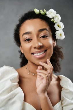 Beyaz bir gelinlik içinde güzel bir Afrikalı Amerikalı gelin saçlarında çiçeklerle gri bir zemine doğru gülümsüyor..