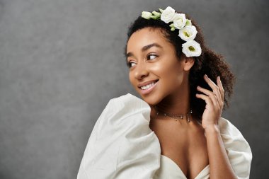 Güzel bir Afrikalı Amerikalı gelin, beyaz bir gelinlik içinde saçlarında çiçeklerle, gri bir arka planda gülümsüyor..