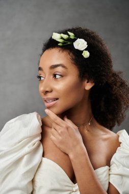 Beyaz bir gelinlik içinde, saçlarında çiçeklerle, gri bir fonda poz veren güzel bir Afro-Amerikalı gelin..