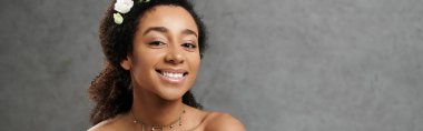 Saçlarında çiçeklerle güzel bir Afro-Amerikalı gelin gri bir arka planda ışıl ışıl gülümsüyor.