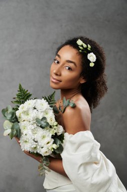 Saçında çiçekler olan beyaz elbiseli Afro-Amerikalı gelin gri arka planda buket tutuyor..