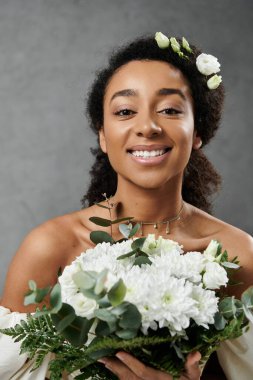 Beyaz gelinlik içinde güzel bir Afro-Amerikan gelinin elinde bir buket beyaz çiçek var, gülüşü gri arka planda neşe saçıyor..
