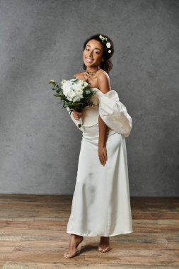 Beyaz gelinlik içinde güzel bir Afrikalı Amerikalı gelin elinde bir buket çiçekle gri bir arka planda dururken gülümsüyor..