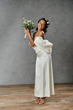 Beyaz gelinlik ve çiçekli taç giymiş güzel bir Afro-Amerikalı gelin elinde buket tutarken ışıl ışıl gülümsüyor..