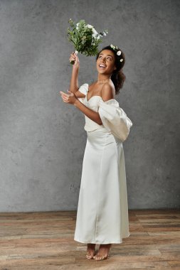 Saçında çiçekler olan beyaz gelinlikli güzel bir Afro-Amerikalı gelin gri bir arka planda duruyor..