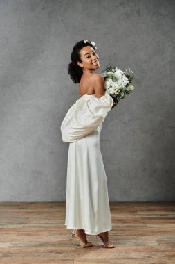 Güzel bir Afrikalı Amerikalı gelin beyaz bir gelinlik içinde saçlarında çiçeklerle gri bir arka planda duruyor..