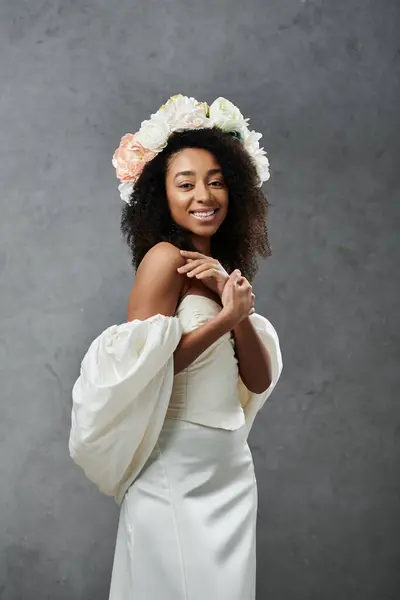 stock image A beautiful African American bride in a white wedding dress and flower crown stands against a grey backdrop, radiating joy and elegance.