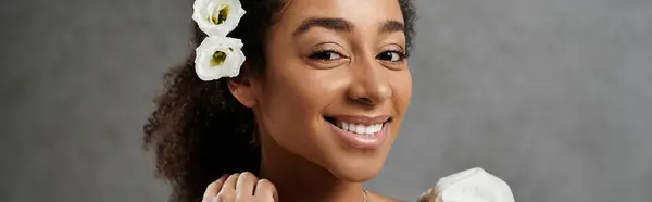 stock image A beautiful African American bride smiles brightly, adorned with flowers in her hair, against a grey background.