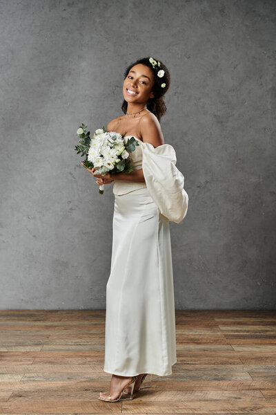 An African American bride in a white wedding dress and floral crown smiles at the camera, holding a bouquet of white flowers.