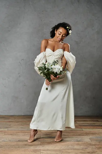 stock image A beautiful African American bride in a white wedding dress holds a bouquet of flowers while standing on a grey background.