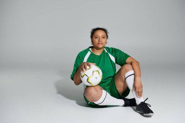 Yeşil futbol forması giyen bayan bir sporcu futbol topuyla oturup, oyuna ara veriyor..