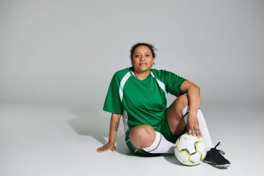 Yeşil futbol formalı bir kadın elinde futbol topuyla beyaz arka planda oturuyor..