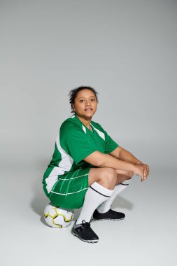 Yeşil futbol formalı kendine güvenen bir kadın stüdyoda bir futbol topunun üzerinde oturuyor..