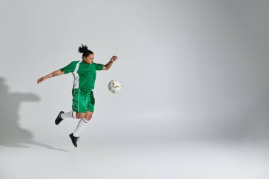 Yeşil formalı ve şortlu bir kadın stüdyo ortamında futbol topuyla hokkabazlık yapıyor..