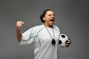 Woman in white jersey raises fist in victory, shouting joyously with soccer ball in hand. clipart