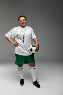 Hakem üniformalı bir kadın, elinde futbol topu tutarak güvenle ayakta duruyor..