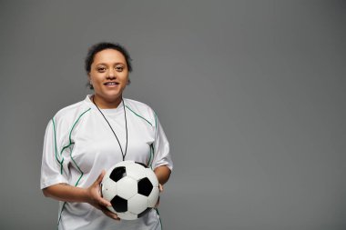 Beyaz formalı atletik bir kadın elinde bir futbol topu tutuyor ve kendinden emin bir şekilde maça hazır gülümsüyor..