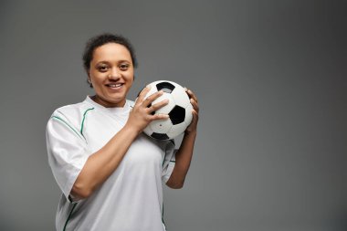 Beyaz formalı genç bir kadın elinde futbol topu tutarken gülümsüyor..