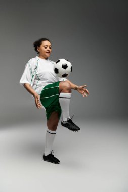 Beyaz formalı ve yeşil şortlu bir kadın stüdyo ortamında futbol topuyla hokkabazlık yapıyor..