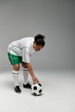 Beyaz formalı ve yeşil şortlu bayan sporcu futbola hazırlanıyor, elini topa uzatıyor.