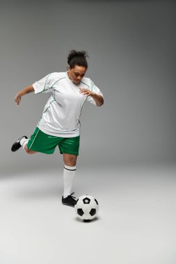 Beyaz formalı ve yeşil şortlu bir kadın stüdyo ortamında futbol topunu tekmeliyor..