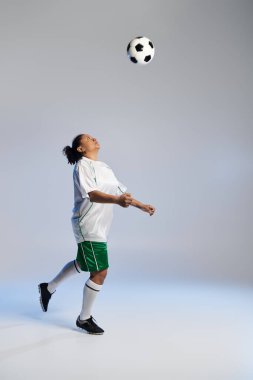 Beyaz formalı ve yeşil şortlu atletik bir kadın stüdyo ortamında futbol topunu yönetiyor..
