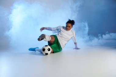 A woman in a white jersey and green shorts plays soccer in a studio setting with a smoky background. clipart